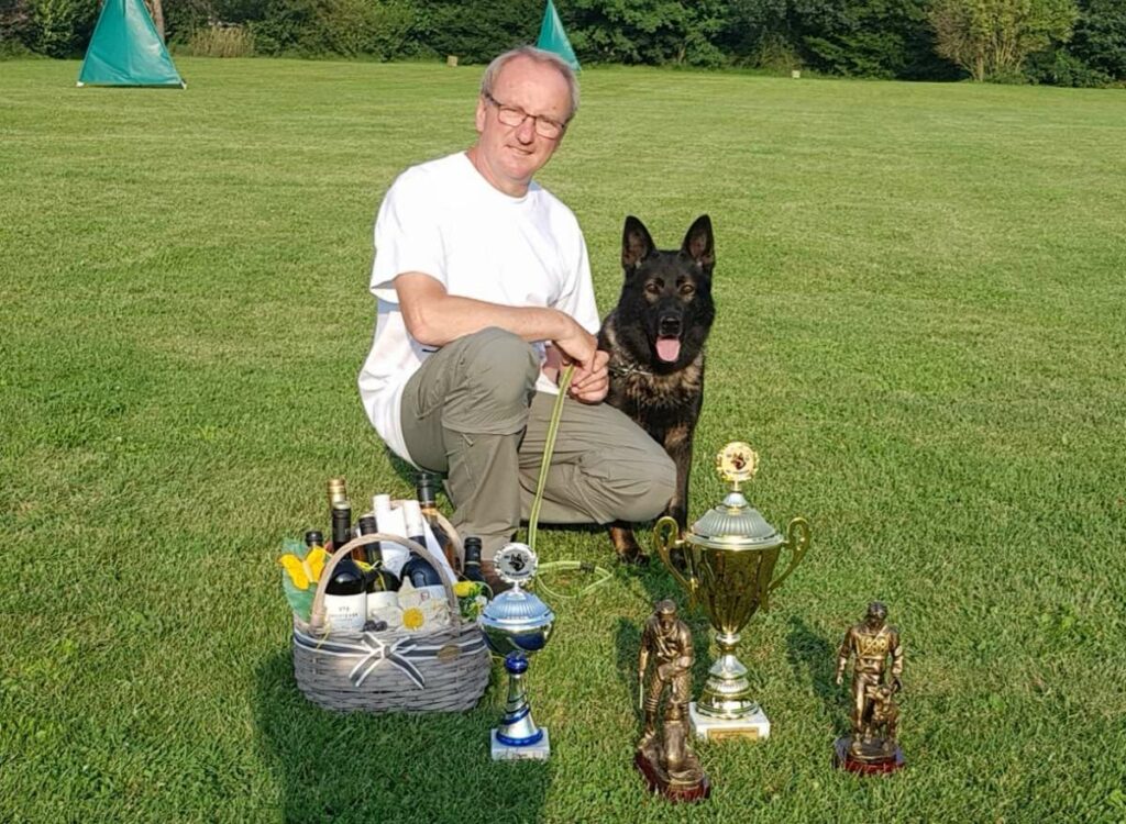 Dieter Barsch mit Pokalen im Vordergrund auf einer Wiese, Schaeferhund daneben